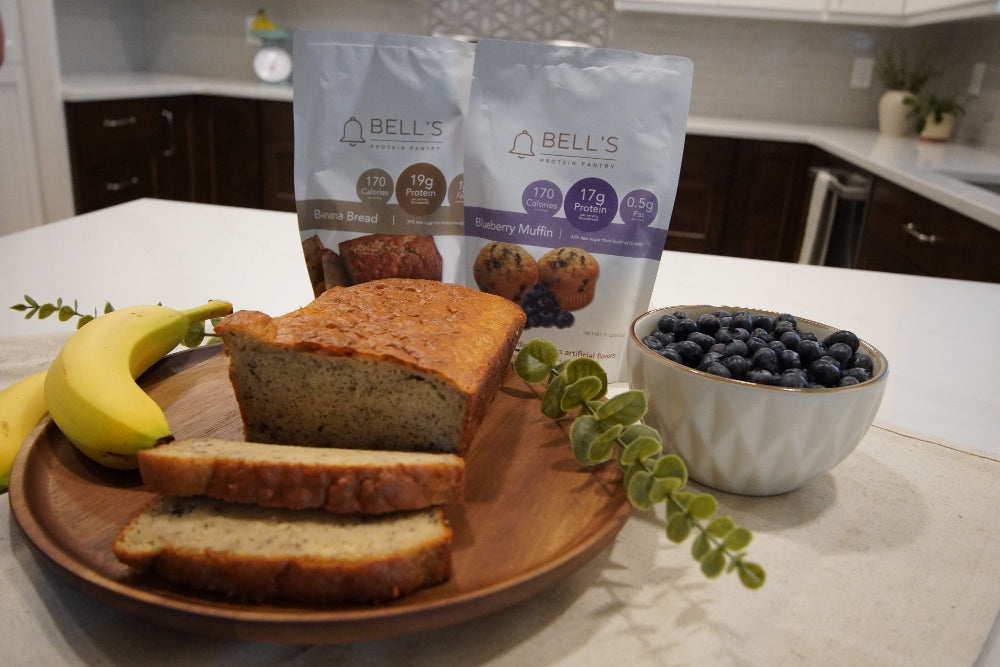 Loaf of Bell's protein banana bread, sliced on a wooden board with bananas and blueberries