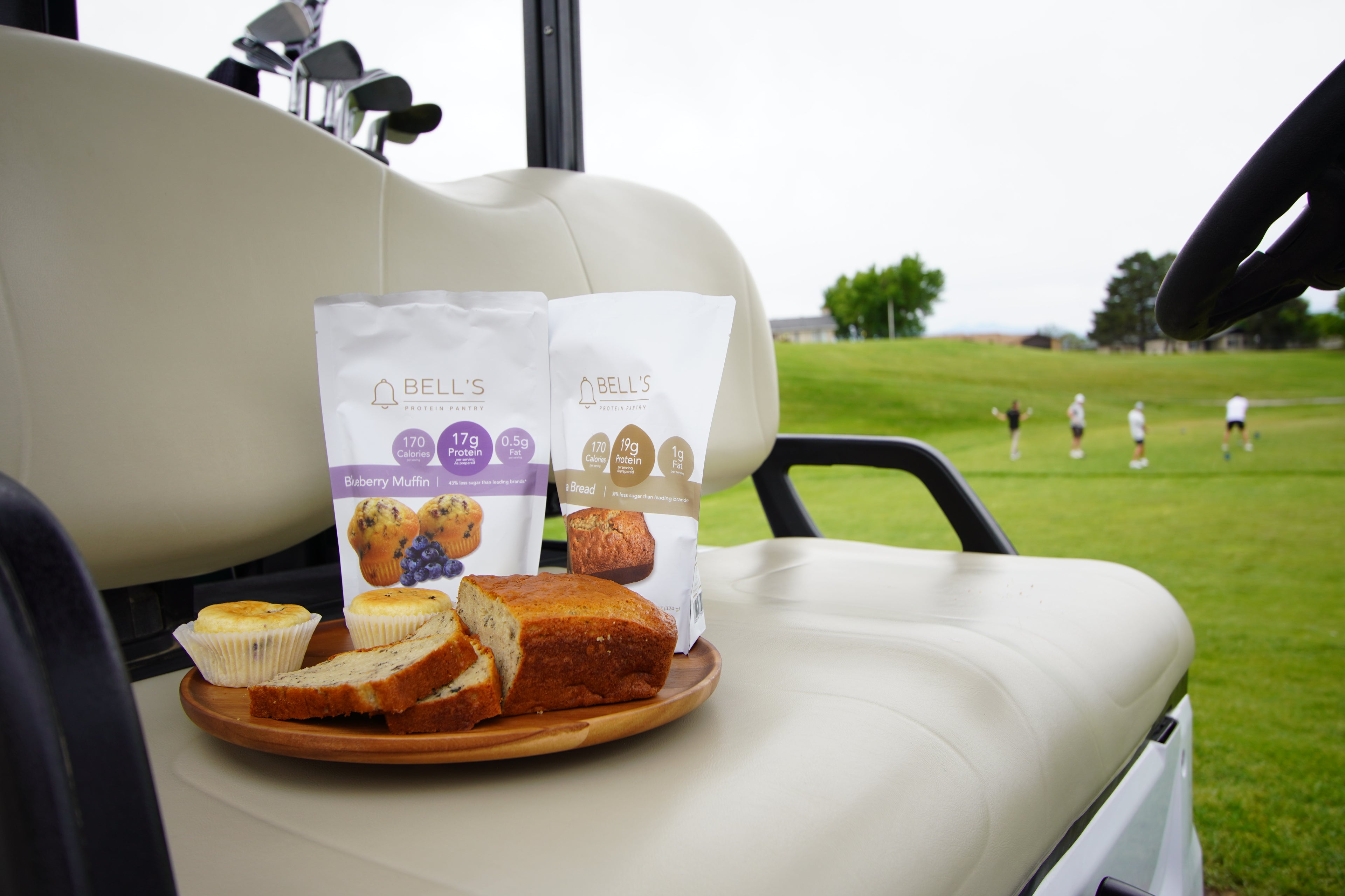 Bread and muffin on a plate with Bell's Protein Pantry packaging on a golf cart seat, golf course in the background