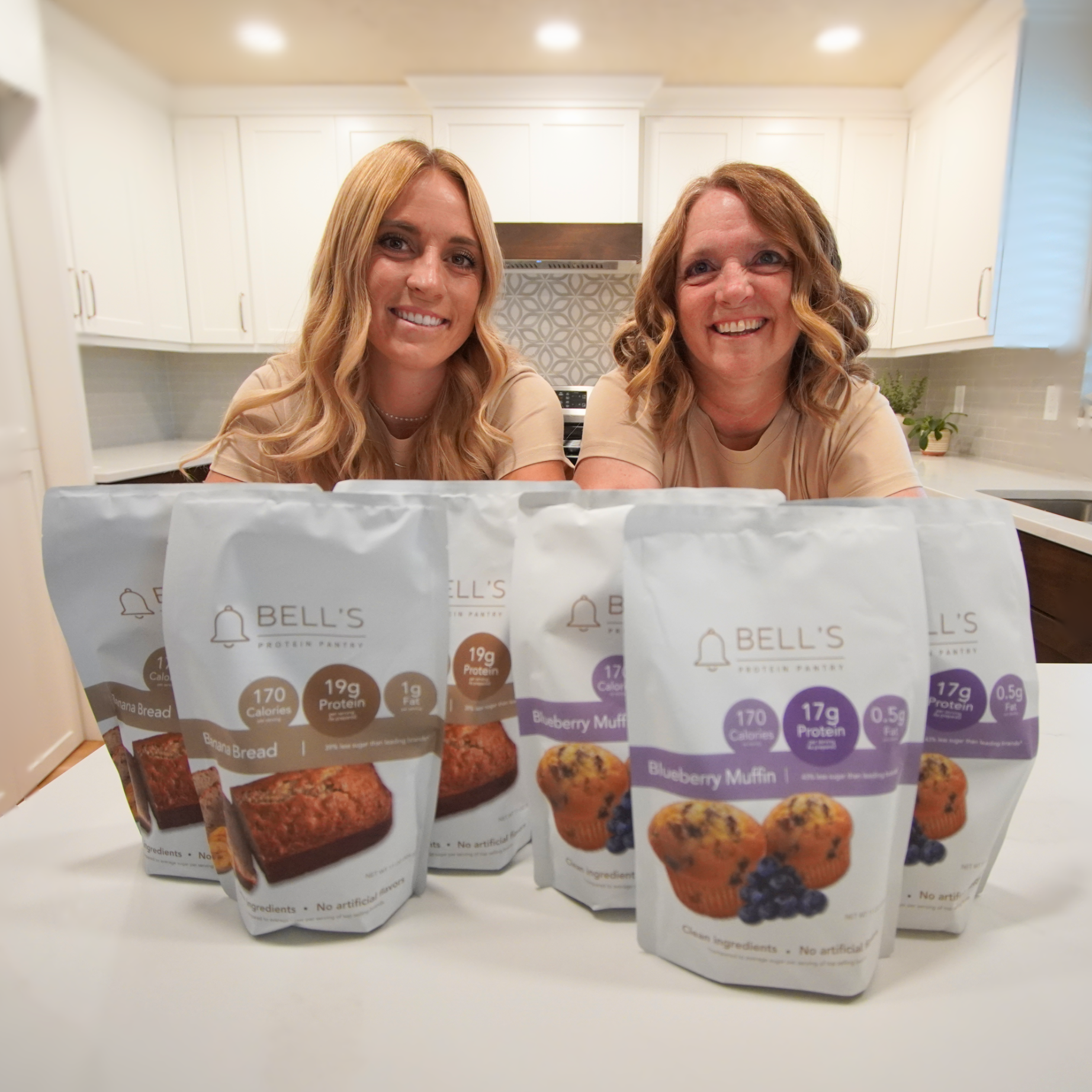 Two owners of Bell's Protein Pantry in a kitchen with packages of Bell's Muffins on a counter.
