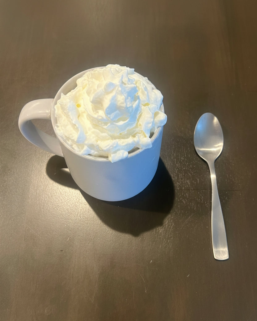 A white ceramic mug filled with whipped cream sits on a dark brown wooden table next to a silver metal spoon.